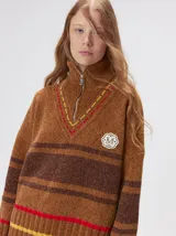 Pullover de punto con textura aterciopelada, diseño oversize, cuello polo con cierre frontal parcial y detalles de pespuntes en contraste. Presenta rayas horizontales en tonos tierra y un parche bordado con la letra M en el pecho.