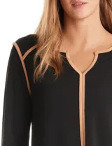 Blusa negra de crepe elastizado con ribetes en contraste color beige que recorren el escote en V, la línea central del frente y las costuras de los hombros. Tiene mangas largas con puños abiertos, abertura lateral y botón metálico.