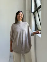 Poncho de tejido de punto color visón, con cuello alto y diseño de trenzas verticales. Presenta mangas cortas ajustadas y un corte holgado y redondeado en el ruedo.
