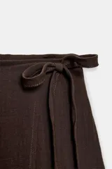 Falda midi pareo de color marrón, con diseño cruzado, cierre lateral con botón interior y lazada. Presenta detalles de bordados y flecos en el bajo.