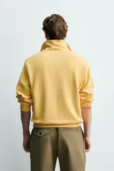 Sudadera de corte relajado confeccionada en algodón con interior de rizo, color amarillo mostaza con efecto lavado. Presenta cuello subido con cierre frontal de cremallera y detalle de cinta trenzada en el tirador, manga larga y acabados en rib.