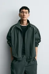 Chaqueta técnica de corte relajado en color verde oscuro, con cuello subido, cierre frontal de cremallera y detalles de líneas en contraste. Presenta bolsillos frontales con cremallera oculta y acabados elásticos en puños y ruedo.