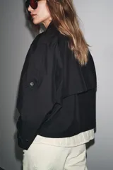 Trench corto negro confeccionado en hilatura con mezcla de algodón. Cuello solapa de muescas ajustable con trabilla y botones. Manga larga. Bolsillos delanteros. Detalle de trabillas en hombros. Cierre frontal cruzado con botones.