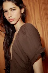 Vestido midi de color marrón, confeccionado en tejido de gasa con textura. Presenta un escote en pico con detalle de lazada frontal, mangas cortas abullonadas con terminación elástica y falda con detalle de godets que aportan volumen. Incluye forro interior a tono y cierre lateral con cremallera oculta.