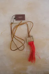 Collar con cordón de cuero tostado y dije geométrico metálico de sección cuadrada con flecos colgantes de hilo rojo.
