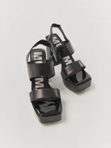 Sandalias de plataforma con taco alto cuadrado y diseño de doble tira frontal. Presentan una puntera cuadrada y cierre con tira elástica en el talón. El diseño combina cuero y acabado en charol brillante.