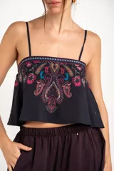 Top corto negro con tirantes finos, escote recto y diseño holgado. Presenta un estampado estilo paisley con detalles en tonos fucsia, turquesa y verde, además de una cenefa geométrica en la parte superior.