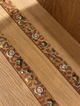 Cinto de cuero con estampado animal print, decorado con tachas metálicas, remaches y ojales plateados. Cuenta con una hebilla metálica cuadrada en tono bronce envejecido.