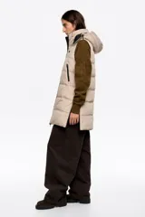 Chaleco acolchado de largo medio y corte en línea A, confeccionado en tejido técnico color beige con detalles en negro. Presenta cuello subido, capucha desmontable, cierre frontal de cremallera, bolsillos laterales con cremallera, logo bordado en el pecho y aberturas laterales con cremallera.