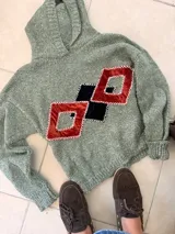 Buzo con capucha tejido en lana merino, color verde jaspeado, con apliques geométricos de terciopelo en tonos terracota y negro, bordados a mano con costuras decorativas en contraste.