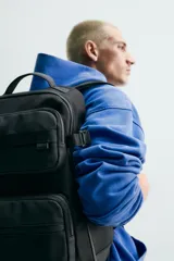 Mochila técnica de color negro con múltiples compartimentos, incluyendo dos bolsillos frontales con cierre, bolsillos laterales y compartimento principal acolchado para dispositivos electrónicos. Cuenta con parte posterior acolchada, banda de sujeción para equipaje, asa de mano y asas de hombro ajustables.