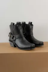 Botinetas de cuero negro con diseño estilo western, punta cuadrada y taco bajo con acabado desgastado. Presentan una tira decorativa con estribo metálico desmontable alrededor del tobillo.
