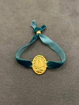 Pulsera con dije ovalado de metal dorado que representa a la Virgen de Fátima, montado sobre una banda de terciopelo elástico en color verde azulado.