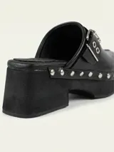 Zueco de cuero encerado color negro, con diseño de punta cuadrada, detalle de tachas metálicas en el lateral y hebilla ajustable en la capellada. Cuenta con suela de goma.