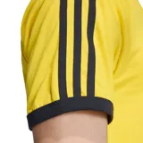 Camiseta Adidas amarilla con cuello redondo y ribetes negros. Presenta las tres rayas características de la marca en los hombros y mangas, y el logo del trébol bordado en el pecho.