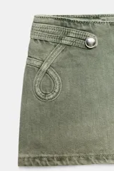 Short mini de tiro bajo en color verde oliva, confeccionado en tejido de algodón con efecto desgastado. Presenta detalles de costuras decorativas, aplicaciones circulares en los laterales y botones metálicos en la cintura. Cuenta con cierre frontal oculto.