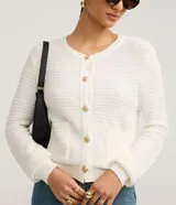 Cardigan de punto texturizado color blanco, con cuello redondo, manga larga y cierre frontal de botones metalizados dorados. Cuenta con dos bolsillos aplicados al frente y puños y dobladillo en rib.