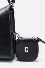 Bolso de hombro tipo baguette en color negro, con acabado brillante y estructura rectangular. Cuenta con una correa combinada de cadena metálica plateada y cuero sintético, y un pequeño monedero extraíble unido a un lateral. Presenta detalles de cordones decorativos en el otro lateral y cierre mediante solapa con imán.