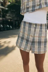 Falda pantalón de tiro alto confeccionada en tejido de tweed con estampado a cuadros en tonos beige, azul y negro. Presenta detalles de falsos botones y cierre lateral con cremallera oculta.