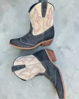 Botas texanas de caña media, confeccionadas en cuero vacuno. Presentan un diseño bicolor que combina el color negro con paneles en color crema, decorados con costuras bordadas en hilo rojo. Cuentan con taco cuadrado de madera y puntera estilizada.