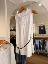 Blusa blanca sin mangas, con cuello trenzado fruncido, detalles de volados en el ruedo y un cinturón fino marrón incorporado.