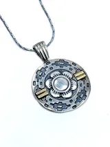 Dije redondo de plata con detalles en oro, con diseño de guarda pampa y flor central.