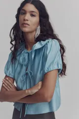 Blusa de viscosa color celeste con cuello Peter Pan fruncido y ribete de encaje. Presenta cierre frontal con tres tiras para anudar, mangas cortas abullonadas y puños con elástico.