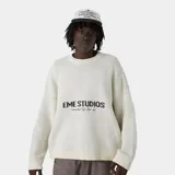 Sweater de punto grueso color blanco hueso, con cuello redondo, corte cuadrado y ligeramente corto. Presenta un gráfico frontal con el texto "EME STUDIOS curated by ben ice" en color marrón. Tiene puños ajustados.