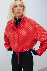 Chaqueta roja con estampado de lunares blancos, cuello subido, manga larga con puños plisados, bolsillos delanteros, bajo ajustable con cordón y cierre frontal con cremallera.