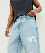 Pantalón baggy de jean celeste con diseño estampado de grafitis en estilo lettering blanco. Cuenta con cintura con trabillas, cierre frontal de botón y cremallera, y bolsillos clásicos.