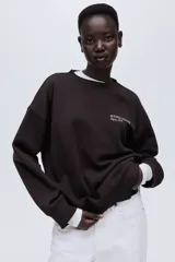 Buzo de tejido de punto color marrón oscuro, con cuello redondo, hombros caídos y puños y bajo acanalados. Presenta un pequeño texto estampado en color blanco en el pecho que dice "RUE DES CAPUCINES Paris, 1991".