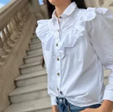 Camisa blanca de algodón con cuello clásico, cierre frontal de botones y detalle de volados en la parte superior delantera y hombros.