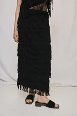 Falda larga negra de silueta recta, con flecos superpuestos en distintos niveles que aportan textura y movimiento.