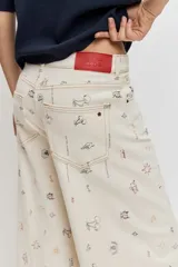 Pantalón de jean wide leg color beige con pequeños bordados y estampados coloridos dispersos, de tiro medio y corte amplio en las piernas.