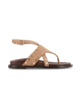 Sandalia plana de cuero gamuza color tostado, con tiras cruzadas estilo ojota adornadas con tachas metálicas doradas y tira ajustable al tobillo con hebilla.