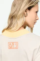 Remera de corte amplio, color beige, con mangas cortas y cuello redondo. Presenta un estampado frontal con la inscripción "The Legendary Hotel Chelsea NY 1884" en tonos celeste y marrón, con detalles en amarillo en el cuello y los puños.