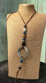 Collar largo tipo gargantilla con tira de cuero marrón, cuentas de madera, piedra y metal, y argollas metálicas colgantes.