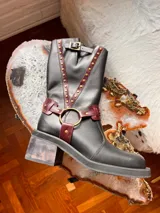 Bota alta de cuero negro con taco cuadrado de efecto madera, puntera redondeada y detalles de tiras en tono bordó con tachas metálicas doradas. Cuenta con una argolla metálica en el empeine y una hebilla de ajuste en la parte superior de la caña.