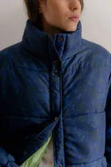 Campera puffer de cuello alto con cierre frontal mediante botones a presión. Presenta un estampado azul oscuro con motivos de paisajes y naturaleza en tonos verdes y azules. El interior está forrado en tela de color verde claro.