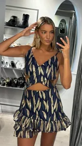 Vestido corto con diseño cut-out en la cintura, tirantes finos y falda con volados en el ruedo. Presenta un estampado de hojas en tonos azul marino y dorado con acabado brillante.