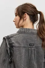 Campera de jean color gris con efecto acid wash, corte clásico y cierre frontal de botones metálicos. Presenta cuello camisero, bolsillos ribeteados y está decorada con apliques de tachas plateadas en el cuello, el canesú frontal, los bordes de la botonera, los bolsillos y el ruedo.