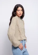 Sweater color crema de tejido trenzado con cuello alto, mangas largas abullonadas y detalle de flecos en el frente.