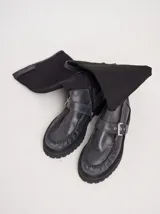 Bota de caña alta con diseño híbrido que combina estilo mocasín y bota. La caña está confeccionada en lona negra, mientras que la parte inferior es de cuero con costuras visibles tipo mocasín. Presenta una hebilla lateral ajustable y suela de plataforma dentada.