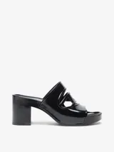 Sandalia tipo mule negra de charol, con plataforma y taco ancho. Presenta un diseño calado en la parte superior con un corte que simula un logo o forma geométrica.