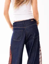 Pantalón de jean estilo wide leg en color azul oscuro, con paneles laterales de encaje en tono marrón rojizo. Presenta un corte amplio y estructura firme sin elasticidad.