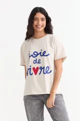 Remera de algodón de corte holgado, color beige, con cuello redondo y mangas cortas con dobladillo. Presenta un estampado frontal con la frase 'joie de vivre' en color azul y un corazón rojo en lugar de la letra 'v'.