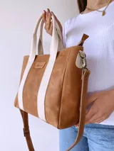 Cartera tipo tote de cuero color camel con detalles de bandas verticales en color crema. Cuenta con doble asa de mano, correa larga ajustable y extraíble, y cierre superior. Incluye etiqueta de cuero con logo grabado en el frente.