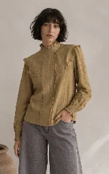 Blusa color mostaza con textura de pequeños puntos y detalles calados verticales. Presenta cuello alto con volados, volados decorativos en los hombros y mangas largas con puños elastizados.