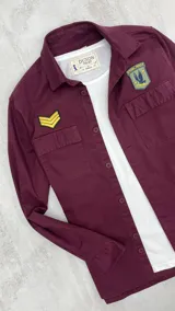 Sobrecamisa bordeaux de gabardina con cuello clásico, cierre frontal abotonado, dos bolsillos con solapa abotonada en el pecho y apliques bordados en el pecho y el hombro. Corte recto y mangas largas con puños abotonados.