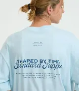 Remera de algodón celeste claro, corte oversized, con cuello redondo y manga corta. Presenta un pequeño texto bordado en azul oscuro en el pecho a cada lado.